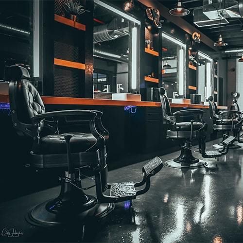 Fotografía en punto de fuga del interior de una barbería. Predominan el color negro y el anaranjado. El piso, la pared y el techo son negros, al igual que las sillas. La barra donde se colocan los objetos y las repisas son anaranjadas. Todo alumbrado con luz blanca. 